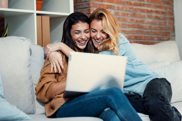 Two people sitting on a couch, smiling and hugging while looking at a laptop.