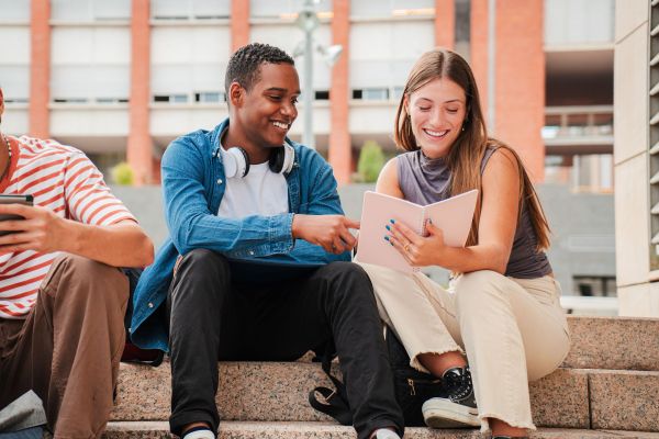 Two people sitting on outdoor steps, smiling and looking at a tablet together.