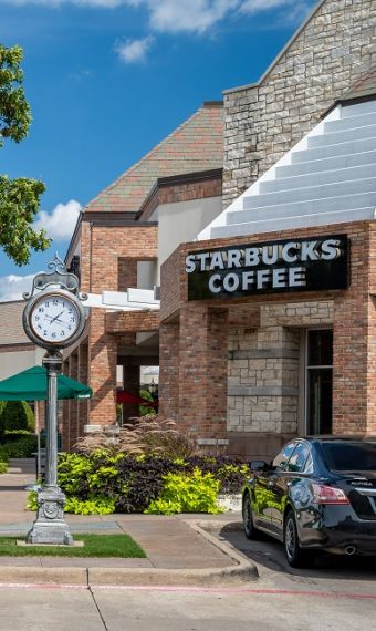 Shopping center with a Starbucks, a clock, parked cars, a motorcycle, and trees under a blue sky.