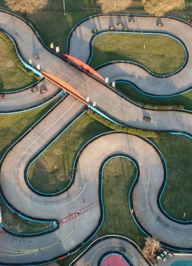 Aerial view of an outdoor go-kart track with winding paths and a bridge. Trees and buildings surround the track.
