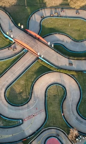 Aerial view of an outdoor go-kart track with winding paths and a bridge. Trees and buildings surround the track.